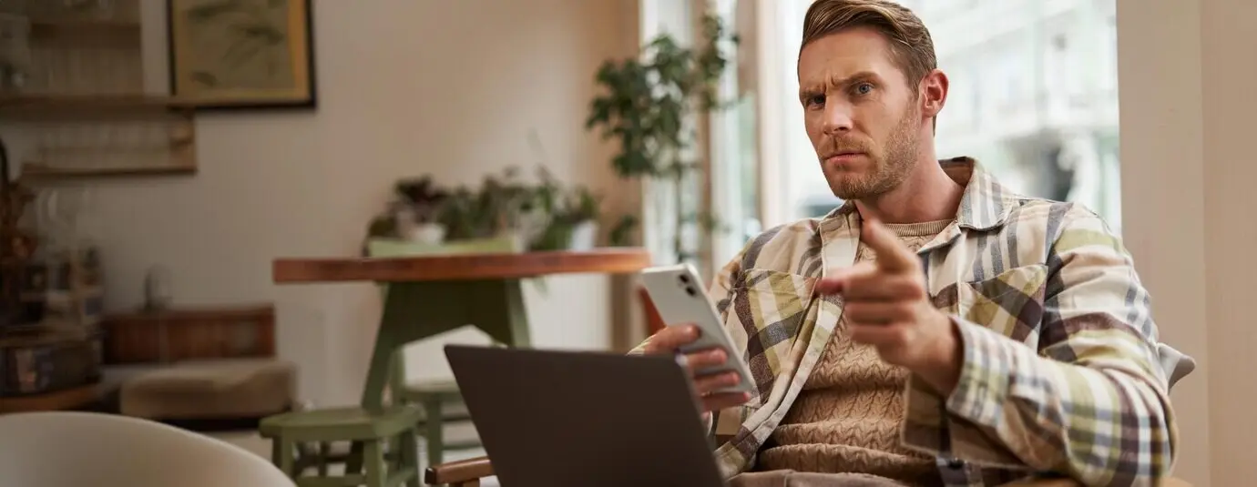 Портрет мужчины с серьезным выражением лица, сидящего в кафе с ноутбуком и смартфоном, предостерегающе указывающего на вас, отчитывающего вас и хмурящегося с неодобрением.