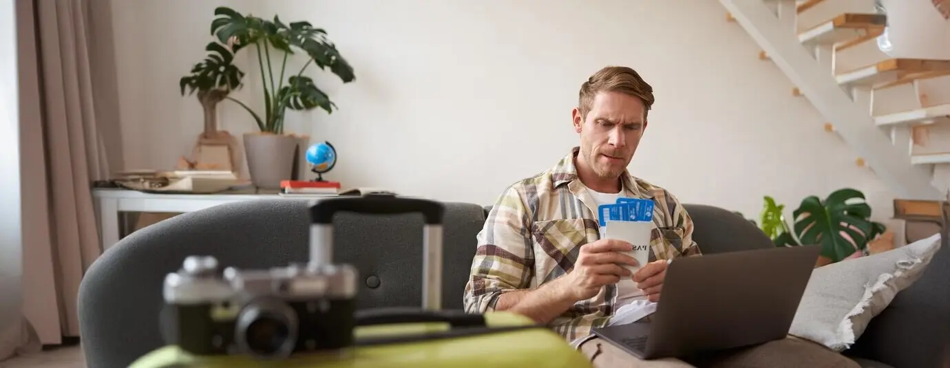 Портрет нахмуренного мужчины, обеспокоенно смотрящего на свои авиабилеты и паспорт; у него открыт ноутбук.